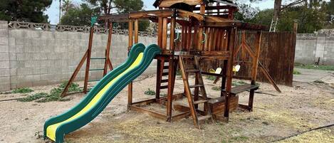 Estructura de madera para juegos infantiles con un tobogán y elementos para escalar, rodeada por un muro de concreto y árboles.