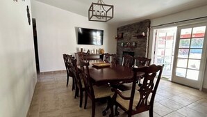 Un espace repas comprenant une grande table et des chaises en bois, un téléviseur à écran plat fixé au mur, une cheminée en pierre et une porte-fenêtre coulissante donnant sur un espace extérieur.
