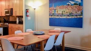 A dining area with a table set for four, a painting of a coastal cityscape on the wall, and a fire extinguisher mounted on the wall. - Sojourn Ocean View Oasis Ocean & Bay Views (Virginia Beach)