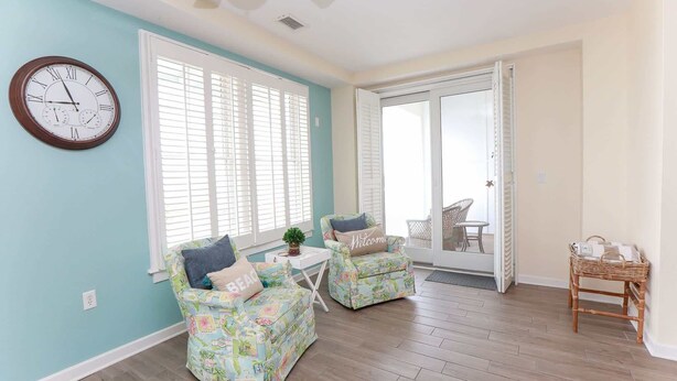 A room with a clock, two chairs, a small table, and a wicker chair. - Sojourn Ocean View Oasis Ocean & Bay Views (Virginia Beach)