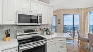 Une cuisine moderne avec des armoires blanches, une cuisinière en acier inoxydable et un four à micro-ondes. On y trouve un coin repas avec une table et des chaises, ainsi qu’un balcon offrant une vue sur l’océan.