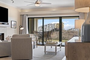 Una sala de estar moderna con chimenea, asientos cómodos, un comedor y un amplio ventanal corredizo que da a un balcón con vistas a un edificio y árboles.
