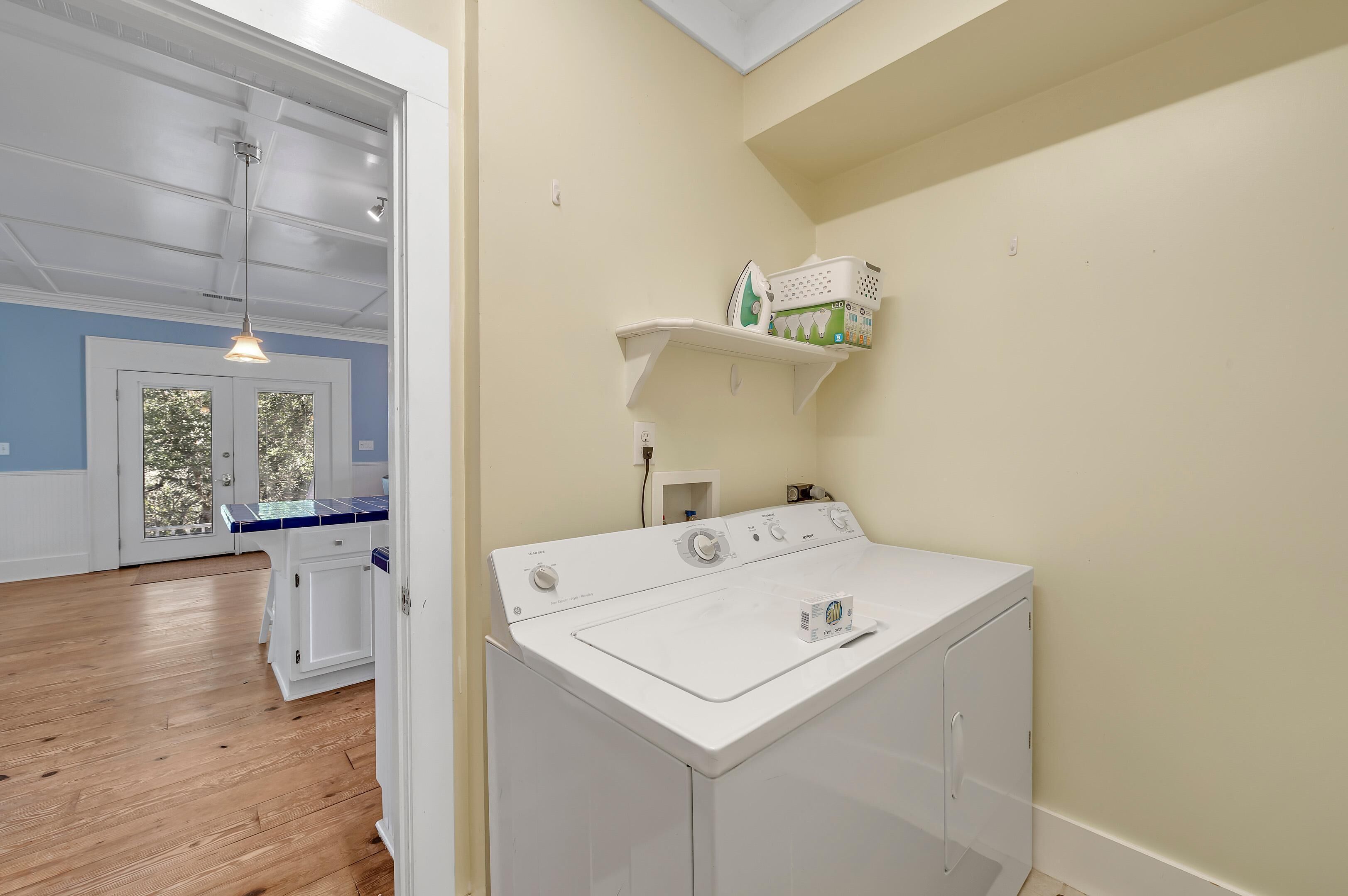 A laundry area with a front-loading washer and dryer, a shelf with laundry supplies, and a view into the adjacent room with a door and a hanging light fixture.
