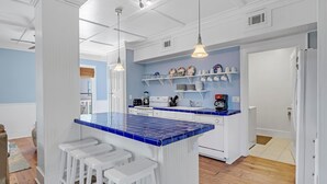 A kitchen with a blue-tiled island, white bar stools, and shelves with dishes and kitchenware. - Ole River Cottage (Pensacola)