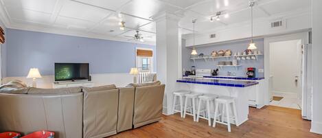 A modern living room with a sectional sofa, a TV, and a kitchen area with bar stools and shelves.