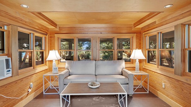 A cozy room with a wooden interior, a sofa, two side tables with lamps, and a large window with a view of trees. - Ole River Cottage (Pensacola)