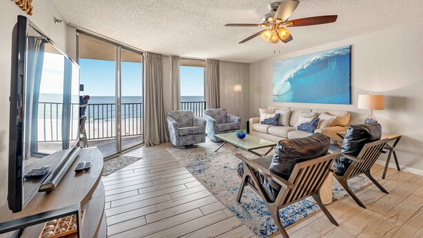 A spacious living room with a balcony, a ceiling fan, a large ocean view painting, and a modern fireplace. - Phoenix East 806 (Orange Beach)