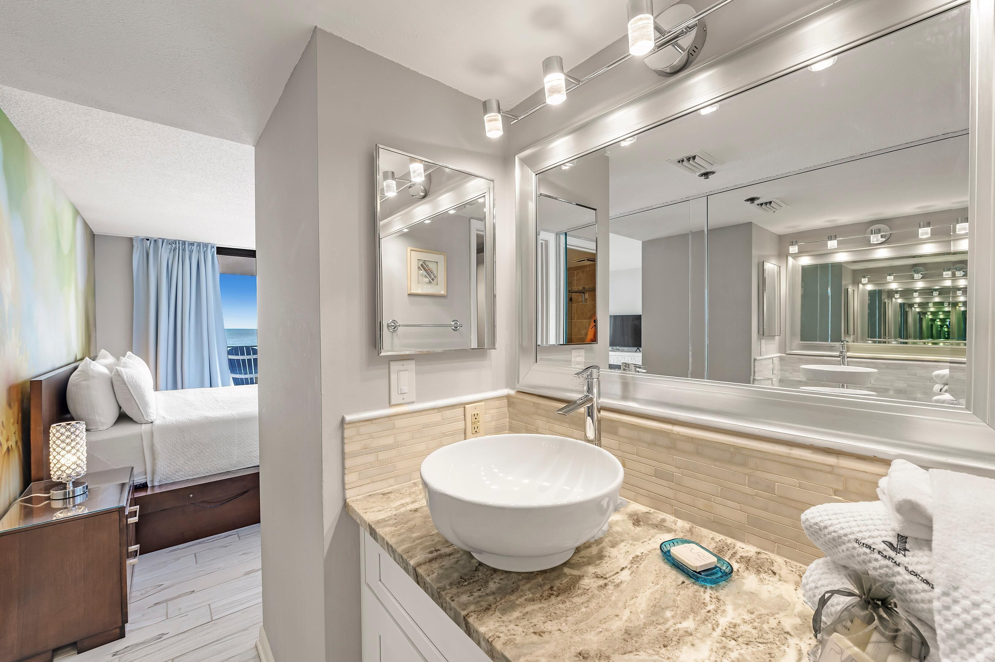 A modern bathroom with a large mirror, a white sink, and a view of the bathroom through the mirror.