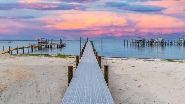 Eine Holzstele führt zu einem Steg, mit freiem Blick auf das Wasser und einem Sonnenuntergangshimmel.