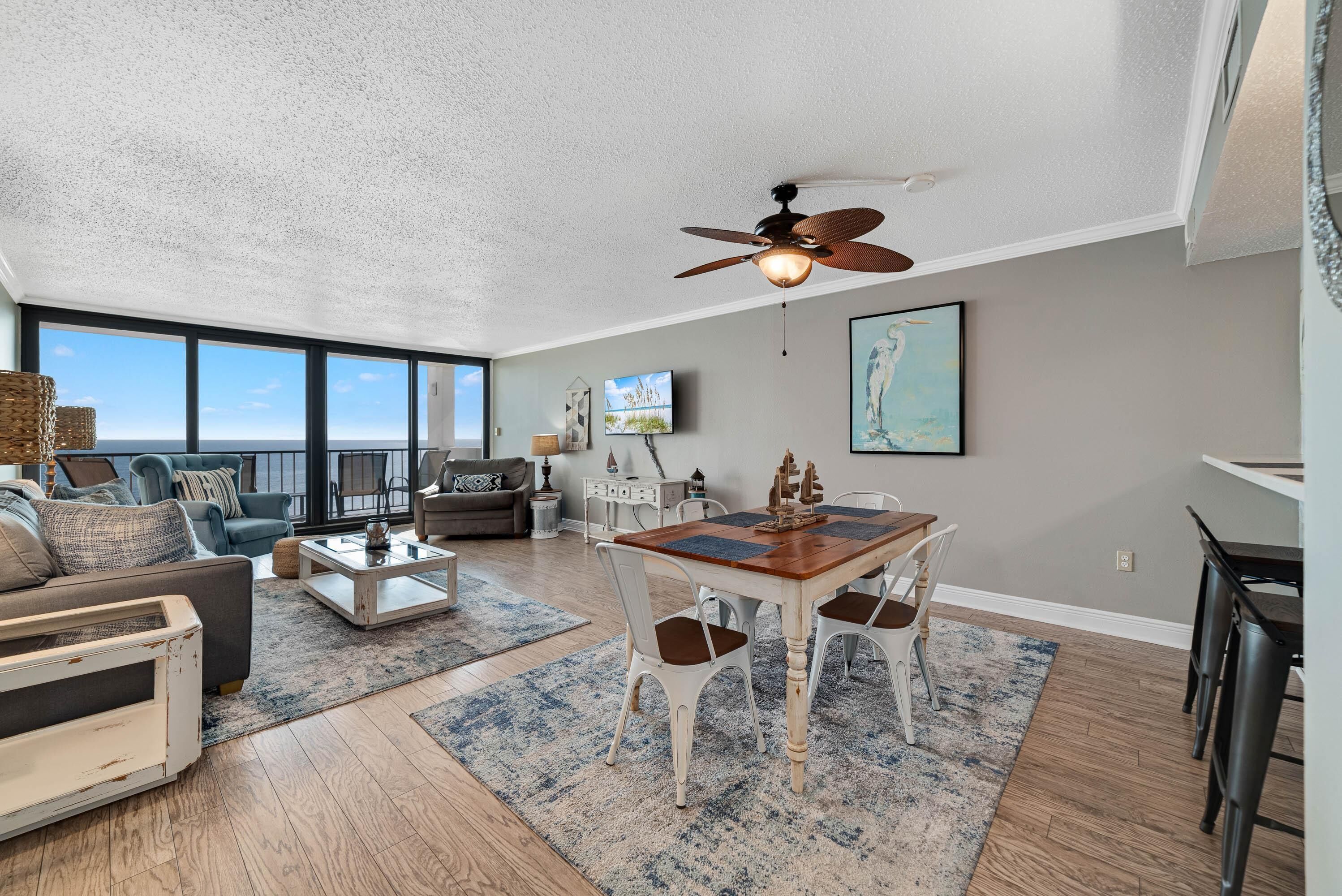 A spacious living room with a large window, a ceiling fan, a dining area with a wooden table and chairs, and a television mounted on the wall.