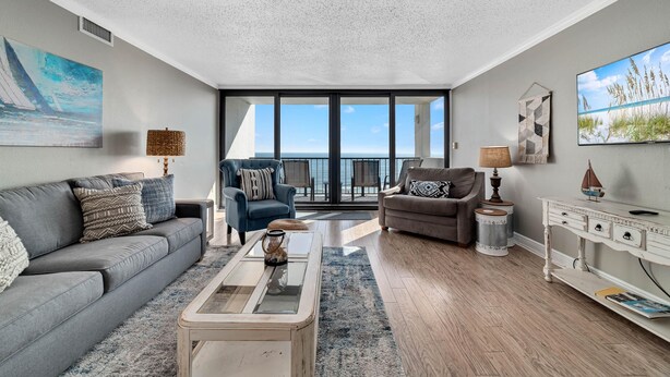 A modern living room with a grey sofa, a blue armchair, and a coffee table. There is a large window with a view of the sea. - The Breakers 61 (Orange Beach)
