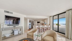 Una sala de estar moderna con televisor, sofá y mesa de centro. Se ve una cocina con comedor al fondo.