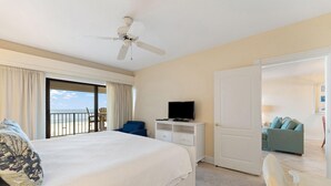 A hotel room with a bed, a television on a white dresser, a balcony with a view of the sea, and a door leading to another room. - Shipwatch A402 (Pensacola)