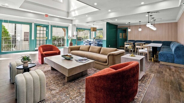A spacious lobby with a high ceiling, a beige sofa, a red armchair, and a blue sofa. There is a small table with a plant and a magazine.