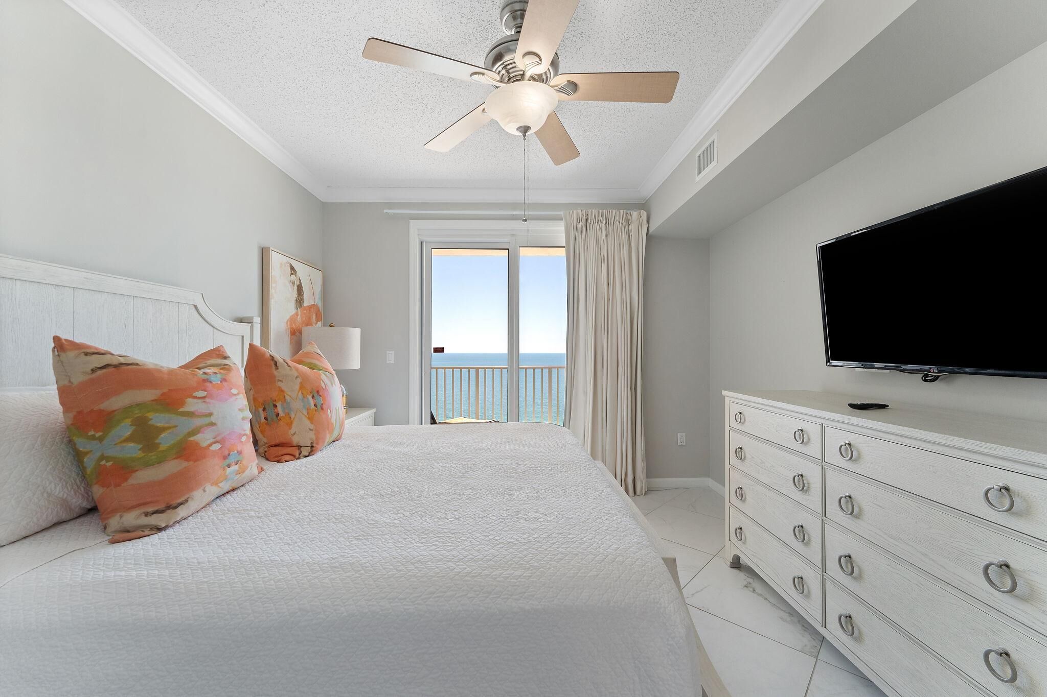 A bedroom with a bed, a dresser, a television, and a view of the sea through a door.
