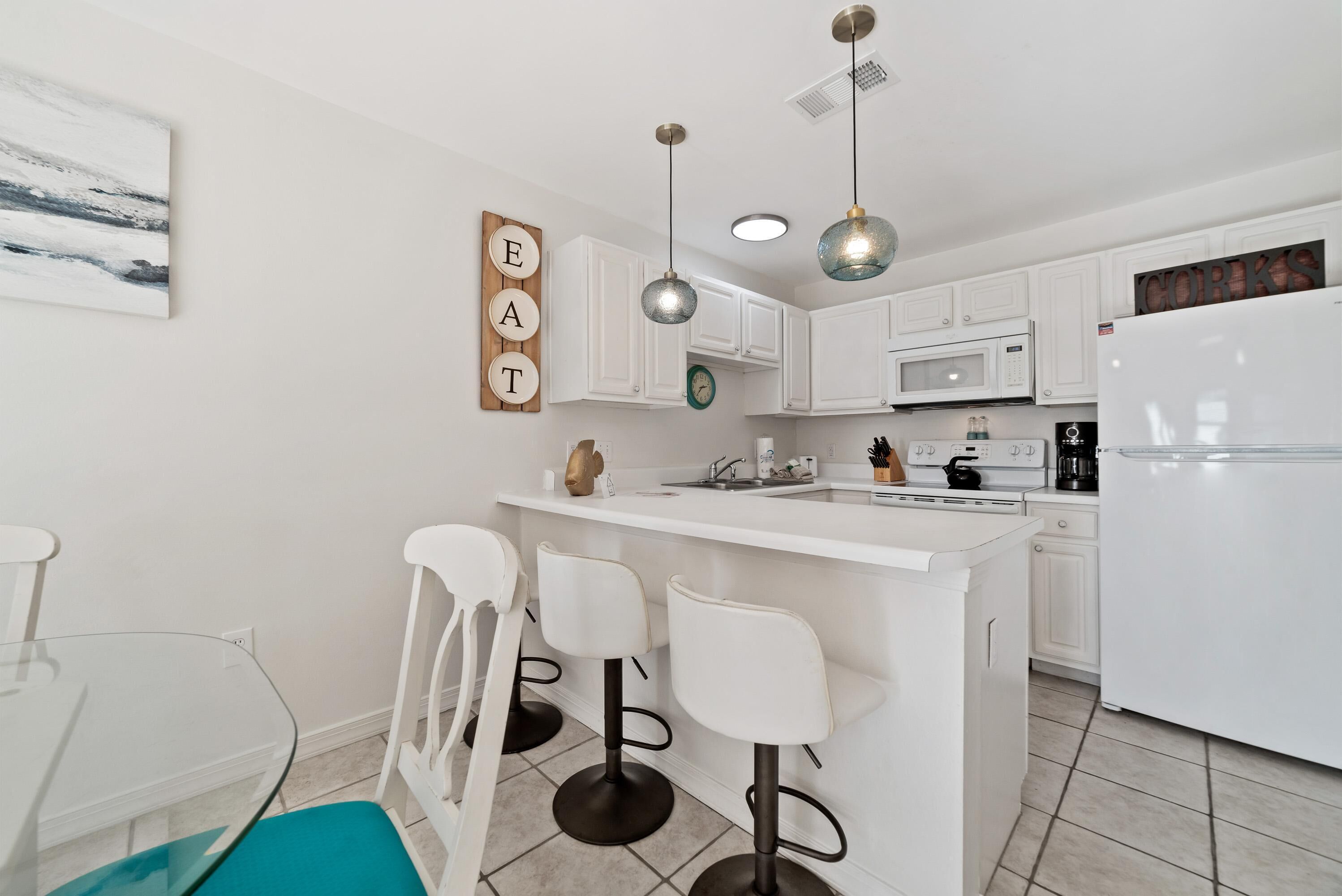 Een moderne keuken met witte kasten, een centraal kookeiland met barkrukken, hanglampen en een witte koelkast.