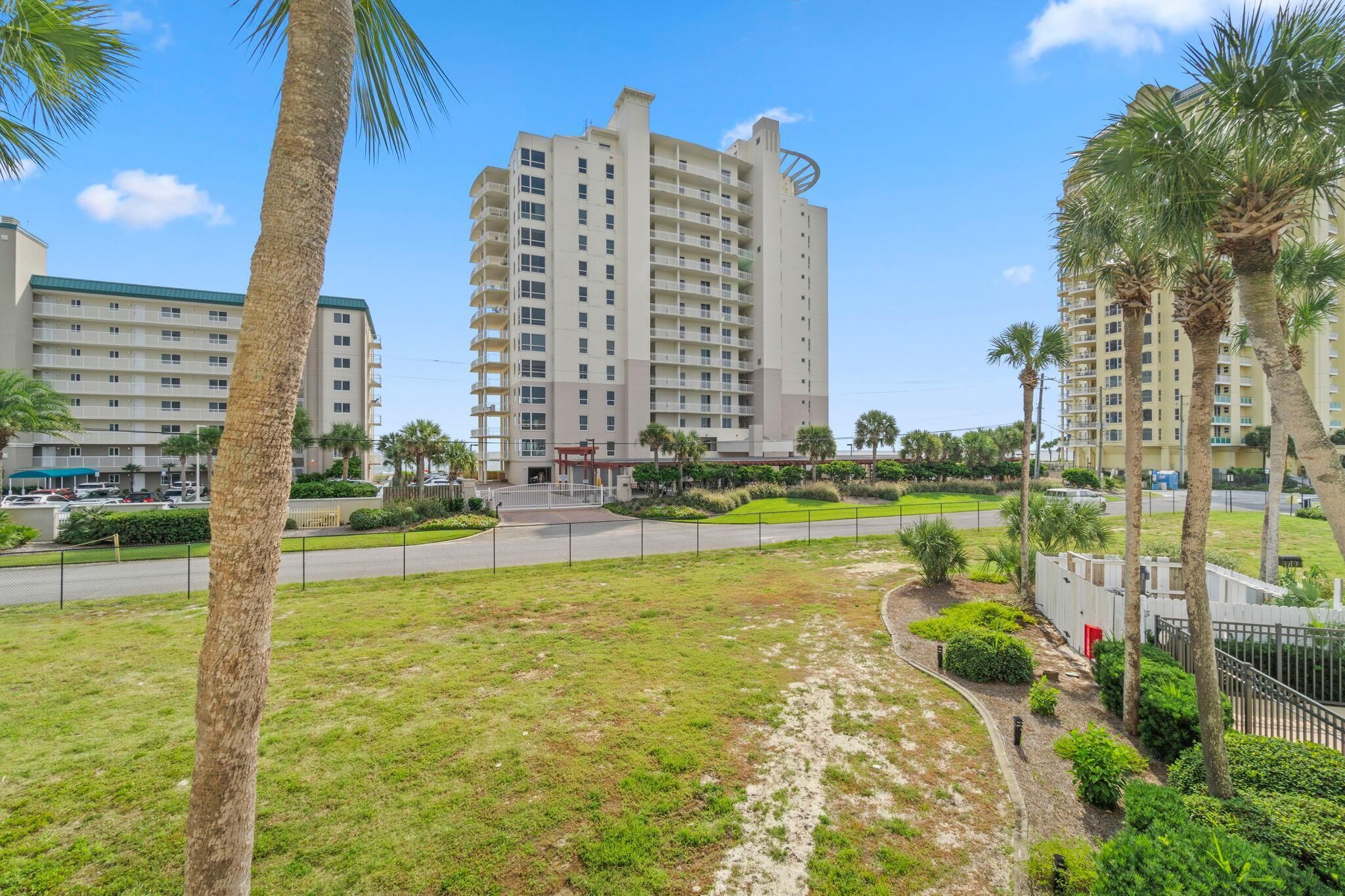 A high-rise hotel with multiple floors, a parking area, and a well-maintained lawn with palm trees.