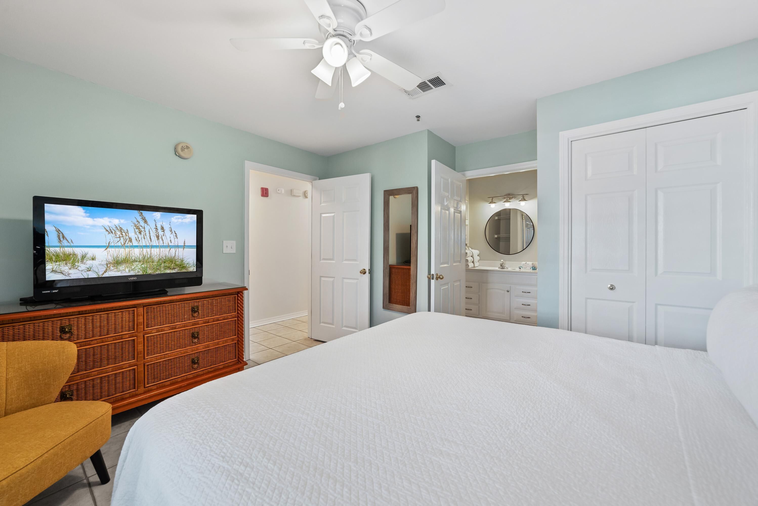 A bedroom with a bed, a television on a dresser, a ceiling fan, and a door leading to a bathroom.