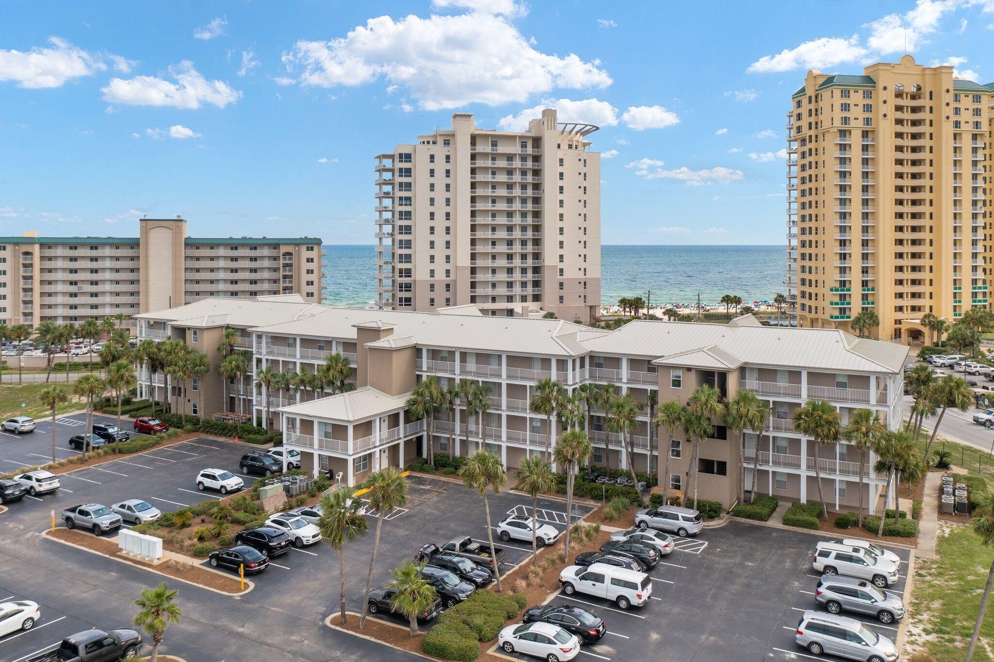 A multi-story hotel with a parking lot, palm trees, and a view of the ocean.