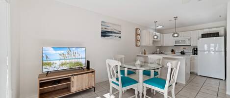 Una cocina moderna con armarios blancos, un refrigerador blanco y un comedor con mesa de vidrio y sillas blancas con cojines color turquesa.