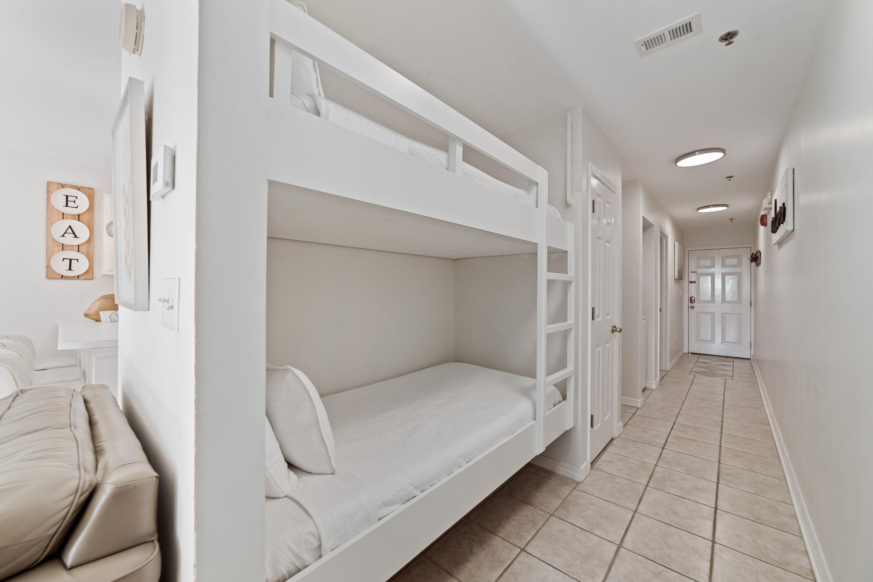 A bunk bed room with a white color scheme, a beige sofa, and a wall-mounted sign that says "EAT".