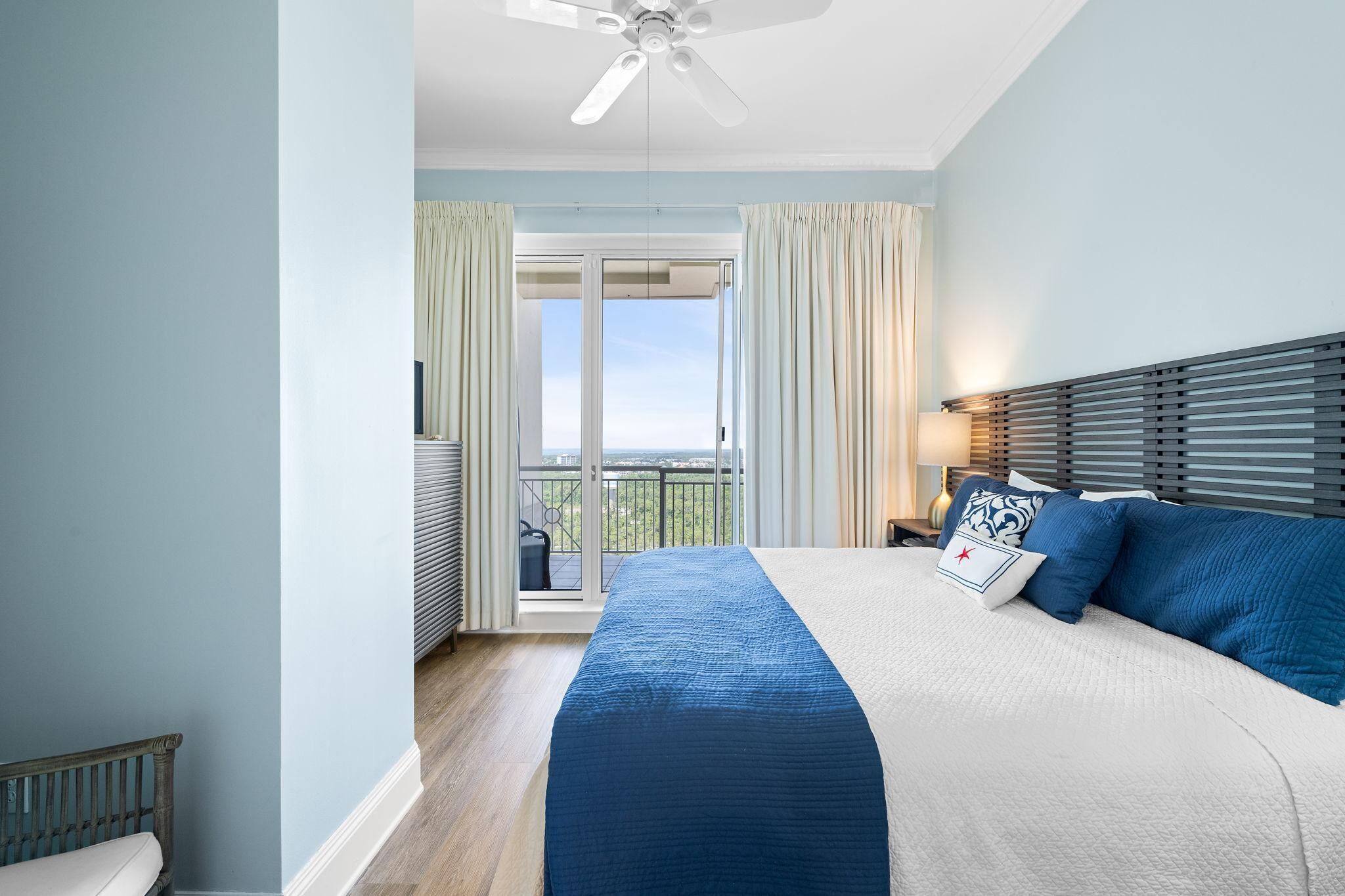 A bedroom with a large bed, a balcony with curtains, a ceiling fan, and a view of the outdoors.