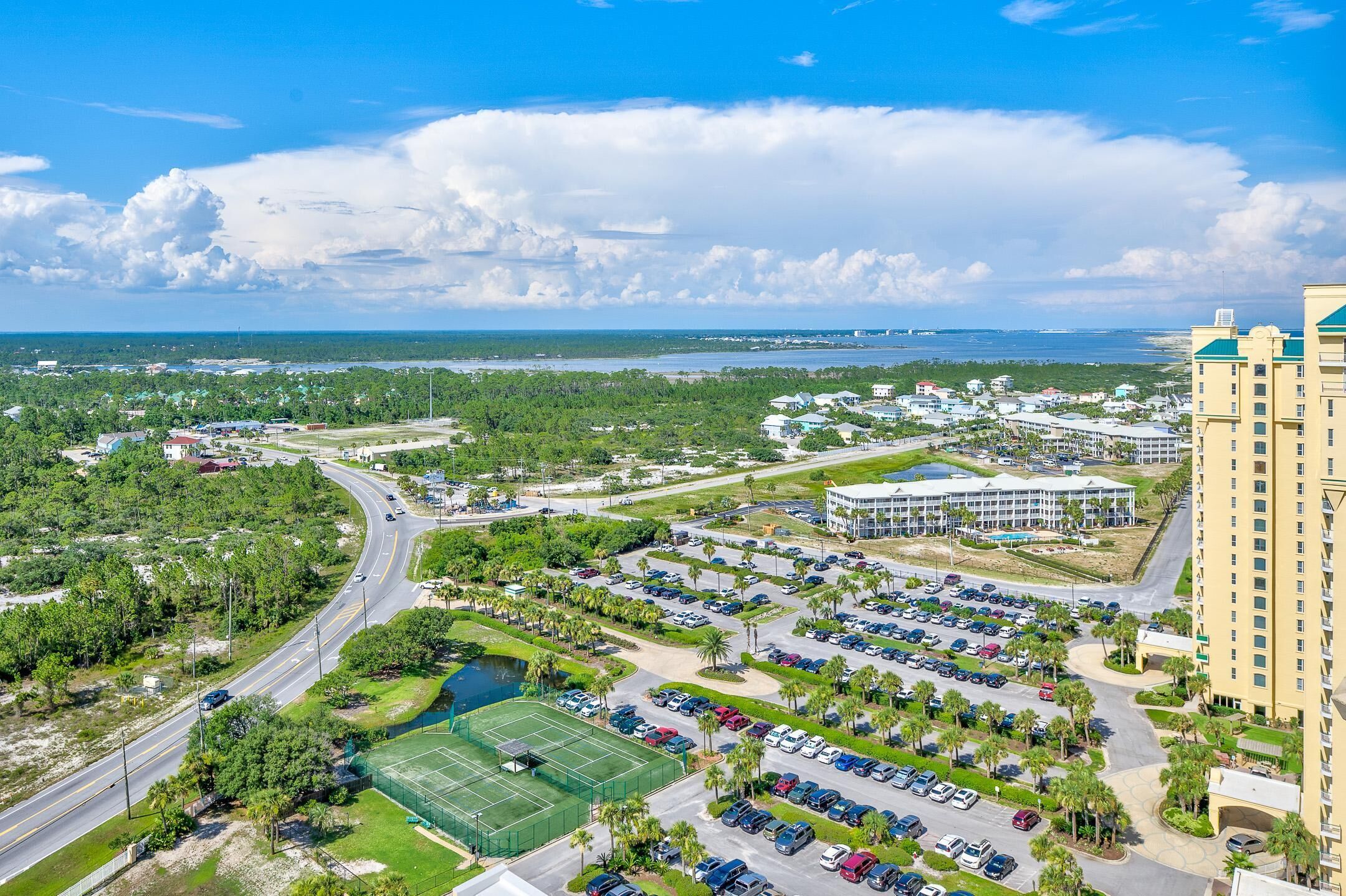 A high-rise hotel with a parking lot, a tennis court, and a view of a coastal area.