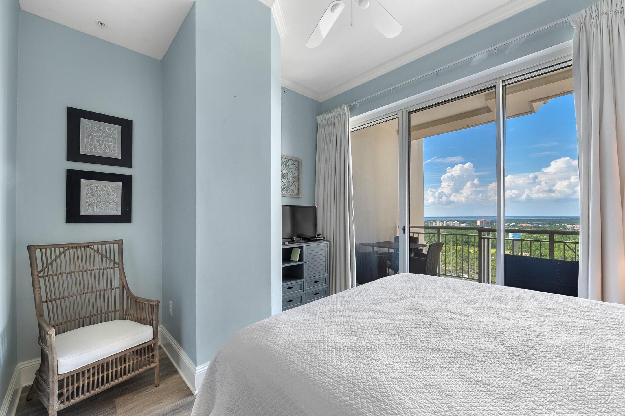 A bedroom with a large bed, a wicker chair, a dresser, and a balcony with a city view.
