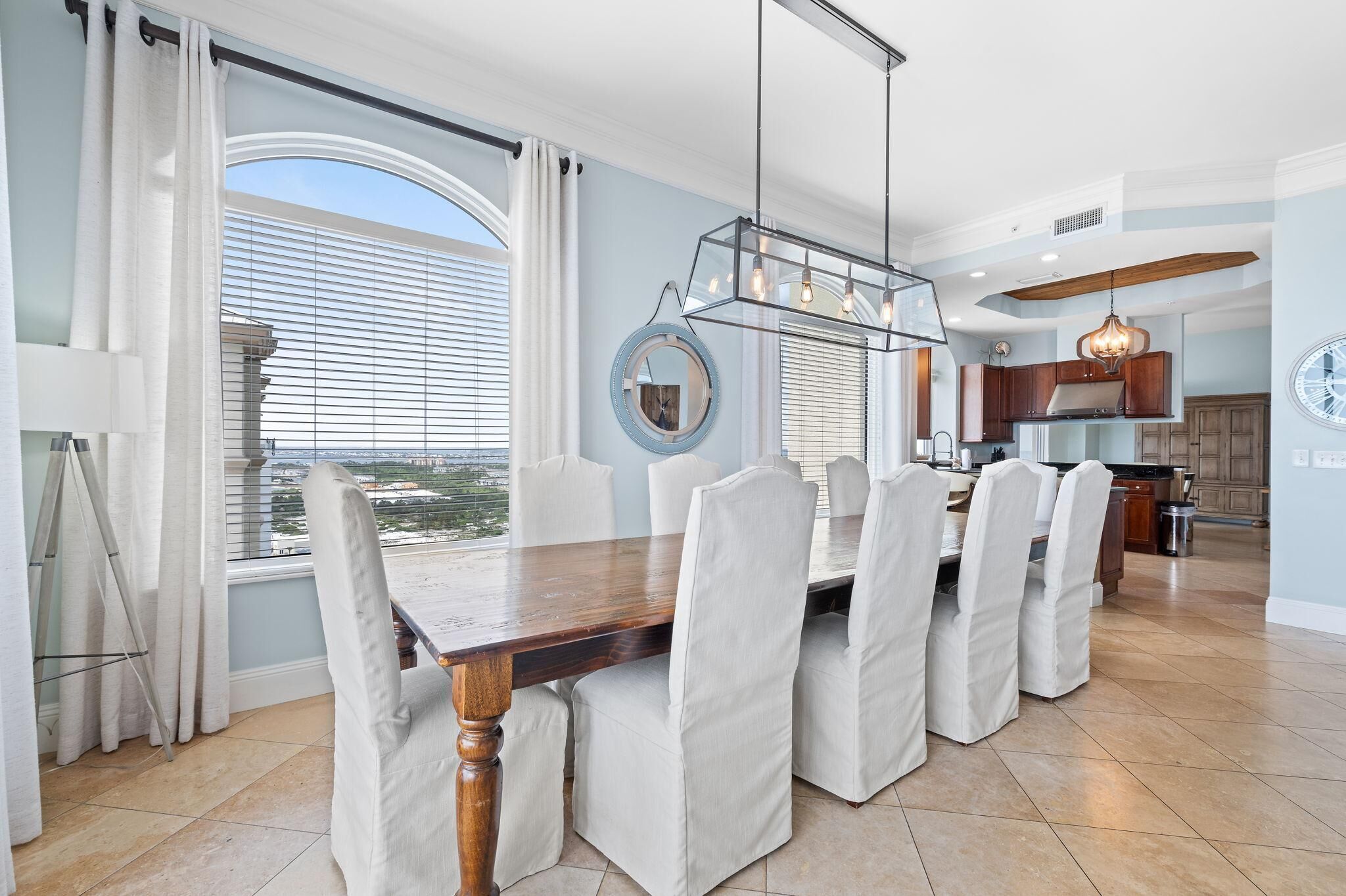 Un coin repas avec une table en bois et des chaises à revêtement blanc, une grande fenêtre avec des stores et, au fond, une cuisine.