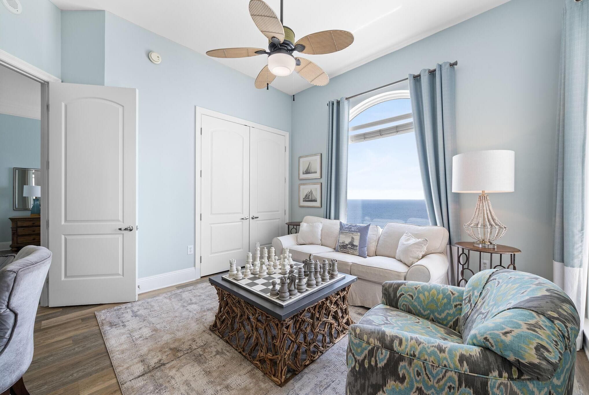 A living room with a chessboard coffee table, a sofa, a patterned armchair, a ceiling fan, and a view of the ocean through a large window.