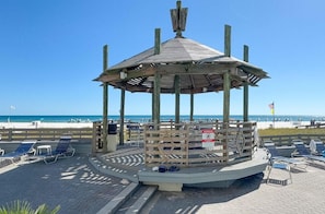 Um gazebo de madeira com telhado, rodeado por espreguiçadeiras e uma placa de aviso, com vista para uma praia.