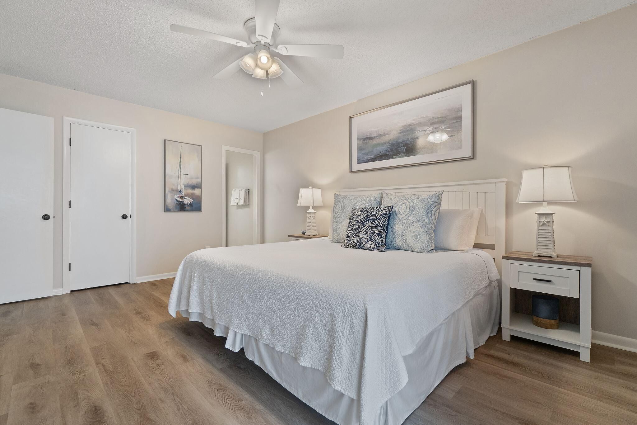 A bedroom with a large bed, a ceiling fan, a nightstand with a lamp, and a framed picture on the wall.