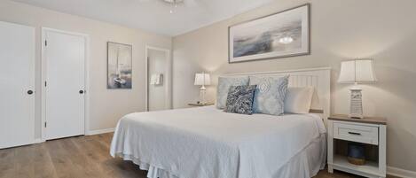 A bedroom with a large bed, a ceiling fan, a nightstand with a lamp, and a framed picture on the wall.