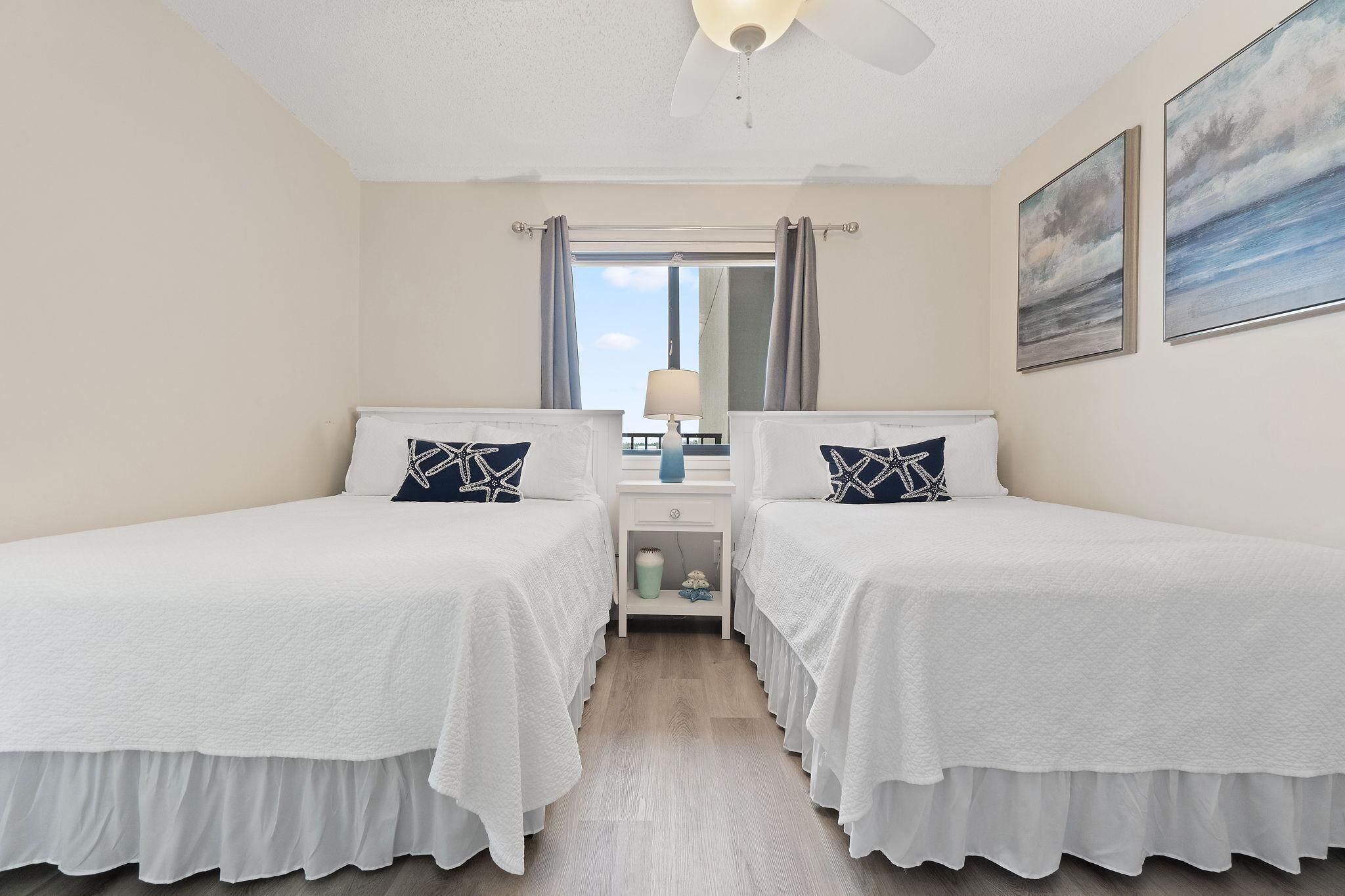 Quarto de hotel com duas camas, uma mesa de cabeceira, um abajur e uma janela com cortinas.