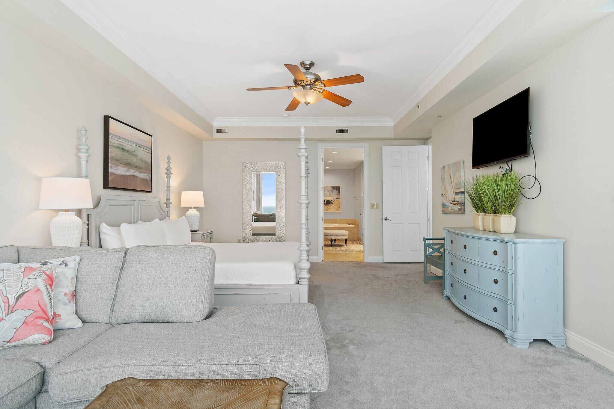 A modern living room with a grey sofa, a wooden coffee table, a ceiling fan, a TV mounted on the wall, and a blue dresser.