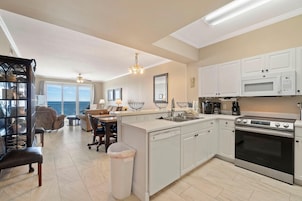 A modern kitchen with white cabinets, a built-in oven, and a dishwasher. There is a dining area with a table and chairs, and a large window offering a view.
