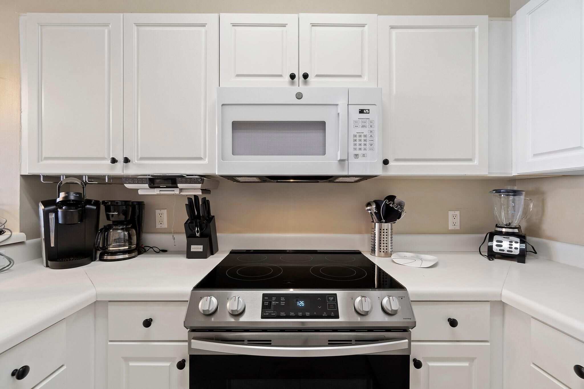 Una cocina moderna con armarios blancos, microondas integrado, cocina con cuatro hornillas y cafetera.