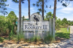 A wooden sign with a bird logo and the text 'The Rookery III' surrounded by trees and a pathway. - Barefoot Bungalow at Rookery (Gulf Shores)