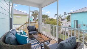 A covered porch with wicker furniture, a glass table, and a view of neighboring houses. - Barefoot Bungalow at Rookery (Gulf Shores)