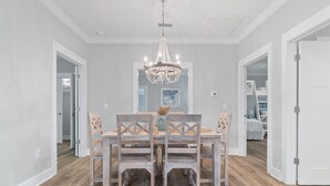 A dining area with a wooden table and chairs, a chandelier, and a view into a bedroom. - Barefoot Bungalow at Rookery (Gulf Shores)