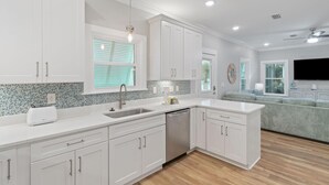 A modern kitchen with white cabinets, a built-in dishwasher, and a large island with a sink. - Barefoot Bungalow at Rookery (Gulf Shores)