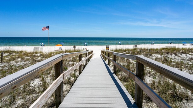 A wooden boardwalk leading to a beach with an American flag, beach umbrellas, and a clear blue sky. - Perdido Sun 502 (Pensacola)