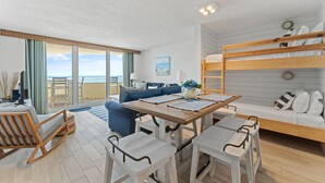 A modern, well-lit room with a wooden bunk bed, a sofa, a dining table with chairs, and a large window overlooking the sea. - Perdido Sun 502 (Pensacola)