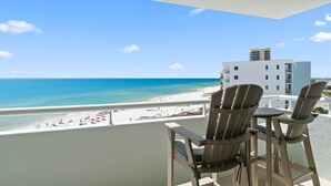 A balcony with two Adirondack chairs and a table overlooking a beach with a white building in the background. - Perdido Sun 706 (Pensacola)