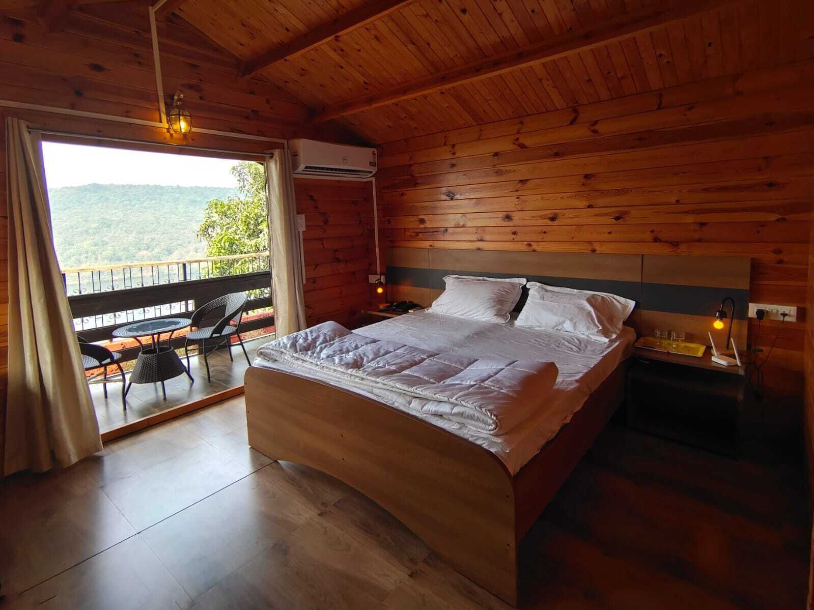A wooden room with a bed, a table with chairs, and a view of greenery outside.