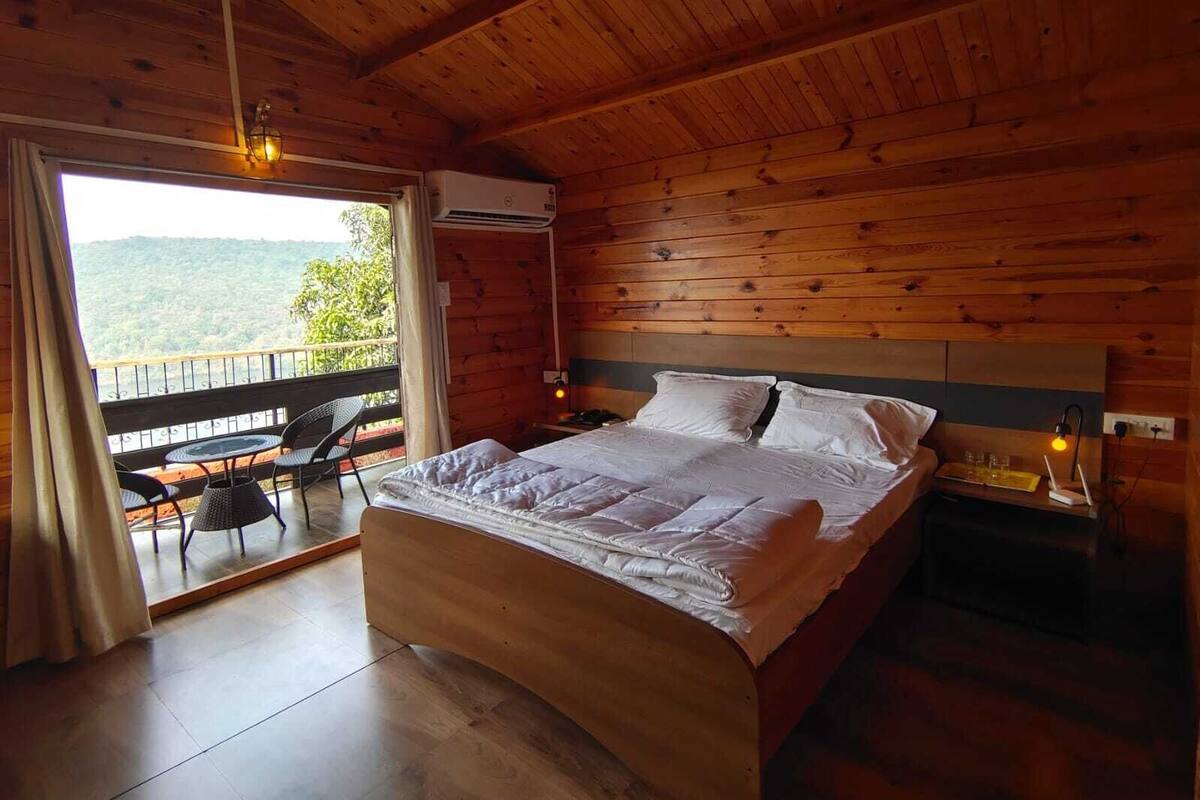 A wooden room with a bed, a table with chairs, and a view of greenery outside.
