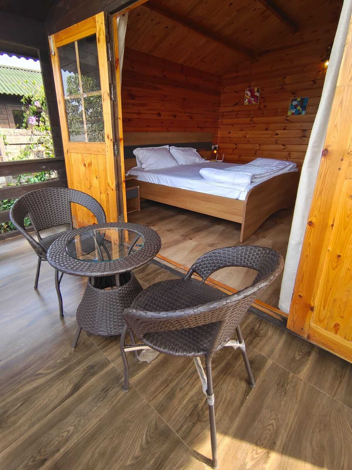 A wooden cabin with a bed, two wicker chairs, and a glass table on a wooden deck.