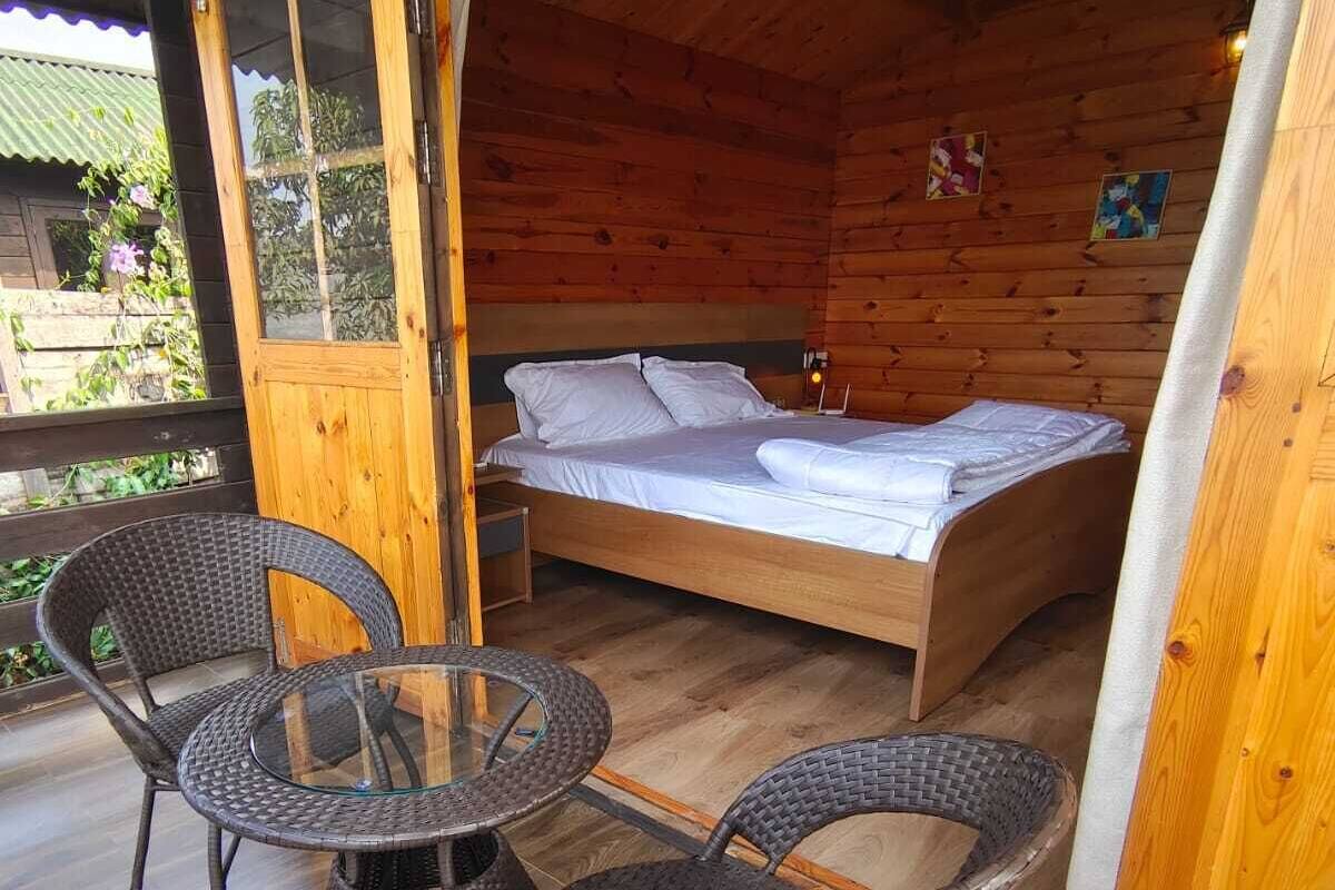 A wooden cabin with a bed, two wicker chairs, and a glass table on a wooden deck.