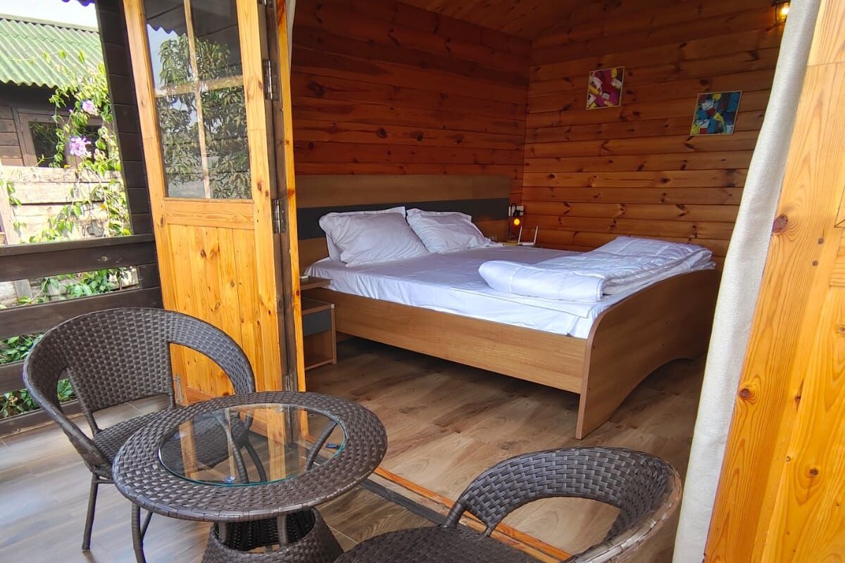 A wooden cabin with a bed, two wicker chairs, and a glass table on a wooden deck.