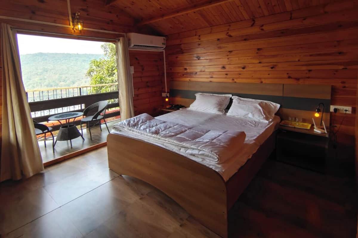 A wooden cabin bedroom with a bed, a balcony with chairs, and a view of the outdoors.